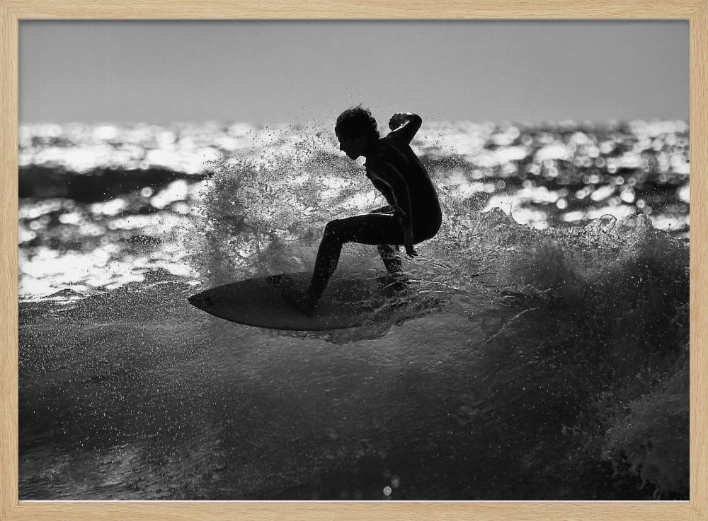 A black and white action shot of a surfer silhouetted against the sparkling ocean. The surfer is crouched low on their board, carving through a wave and creating a dramatic spray of water. The photo is enclosed in a silver frame. Print