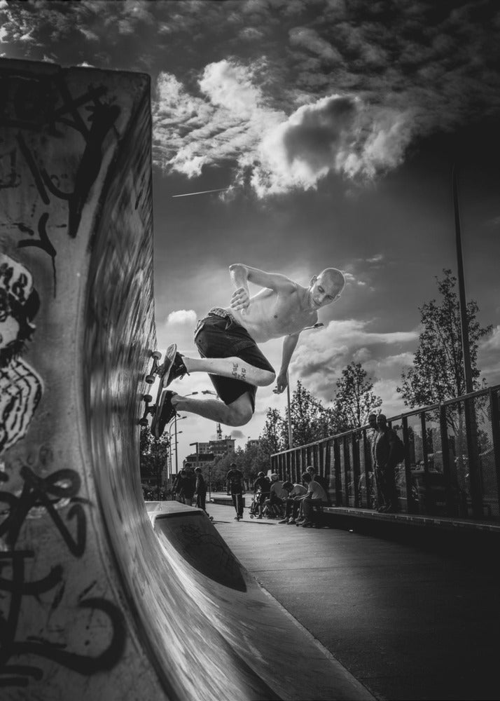 A dramatic black and white, low-angle action shot of a shirtless skateboarder suspended in mid-air at the peak of a graffiti-covered skate ramp, against a backdrop of a cloudy sky. Decor
