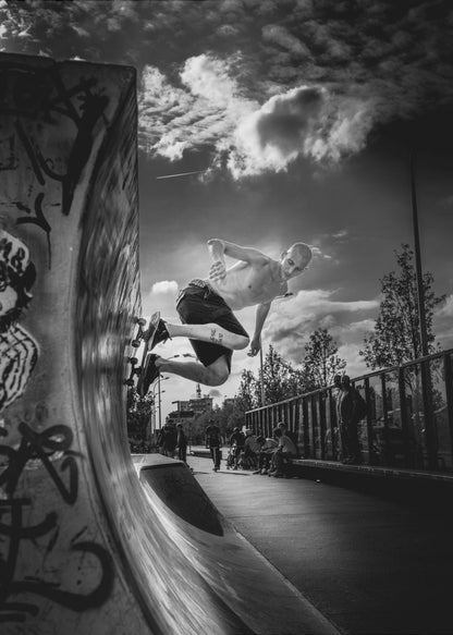 A dramatic black and white, low-angle action shot of a shirtless skateboarder suspended in mid-air at the peak of a graffiti-covered skate ramp, against a backdrop of a cloudy sky. Decor