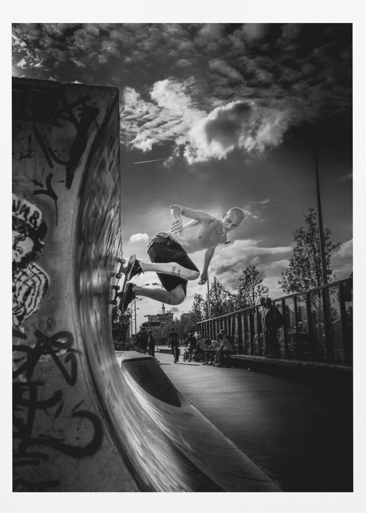 A dramatic black and white, low-angle action shot of a shirtless skateboarder suspended in mid-air at the peak of a graffiti-covered skate ramp, against a backdrop of a cloudy sky. Decor