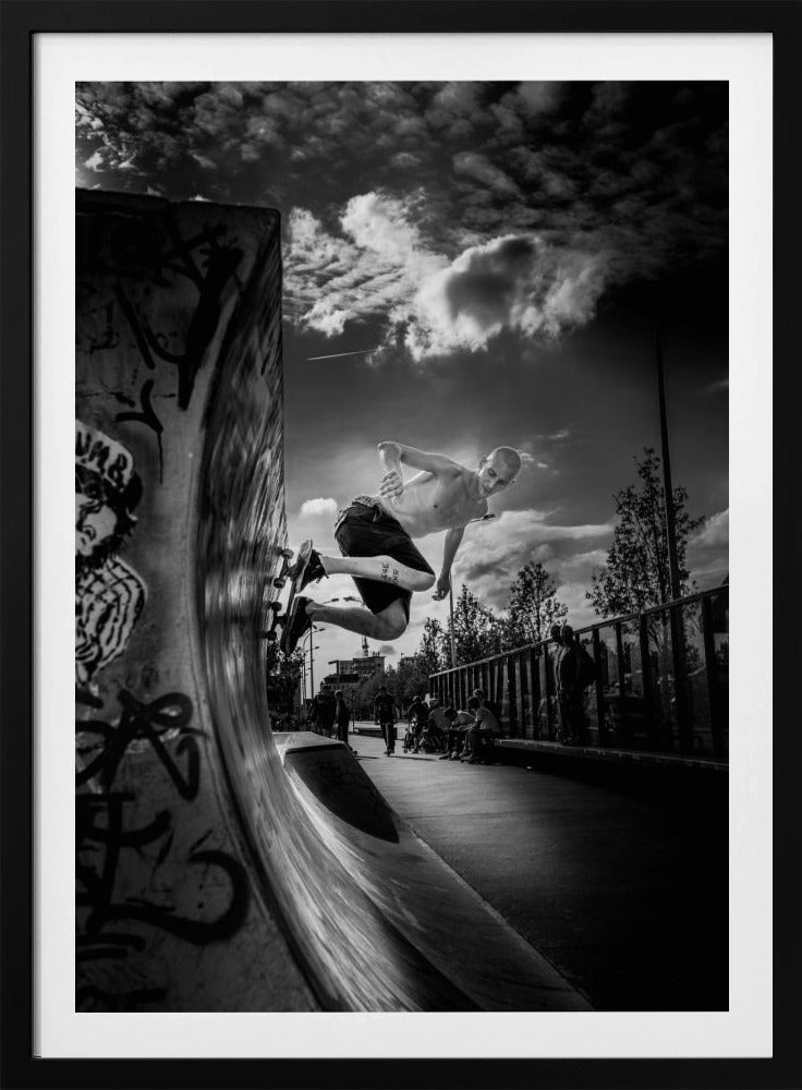 A dramatic black and white, low-angle action shot of a shirtless skateboarder suspended in mid-air at the peak of a graffiti-covered skate ramp, against a backdrop of a cloudy sky. Decor