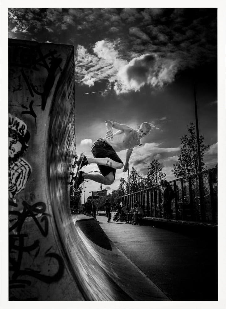 A dramatic black and white, low-angle action shot of a shirtless skateboarder suspended in mid-air at the peak of a graffiti-covered skate ramp, against a backdrop of a cloudy sky. Decor