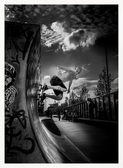 A dramatic black and white, low-angle action shot of a shirtless skateboarder suspended in mid-air at the peak of a graffiti-covered skate ramp, against a backdrop of a cloudy sky. Decor