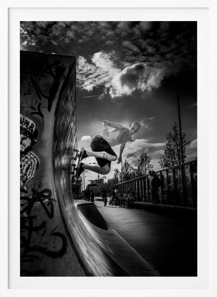 A dramatic black and white, low-angle action shot of a shirtless skateboarder suspended in mid-air at the peak of a graffiti-covered skate ramp, against a backdrop of a cloudy sky. Decor