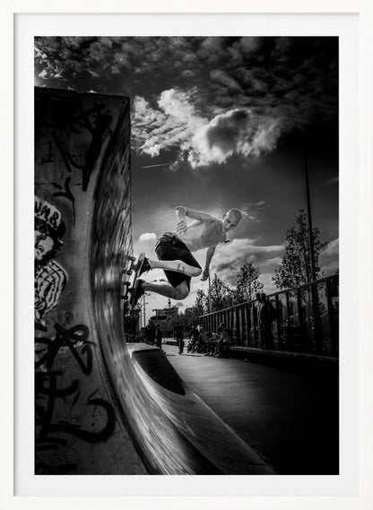 A dramatic black and white, low-angle action shot of a shirtless skateboarder suspended in mid-air at the peak of a graffiti-covered skate ramp, against a backdrop of a cloudy sky. Decor
