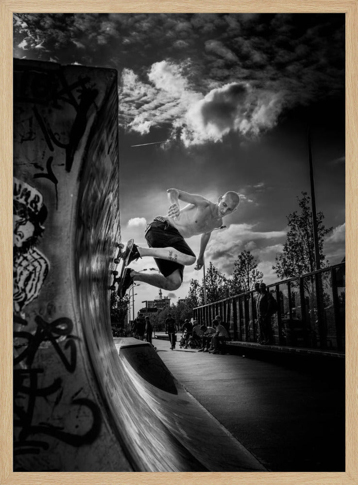 A dramatic black and white, low-angle action shot of a shirtless skateboarder suspended in mid-air at the peak of a graffiti-covered skate ramp, against a backdrop of a cloudy sky. Decor