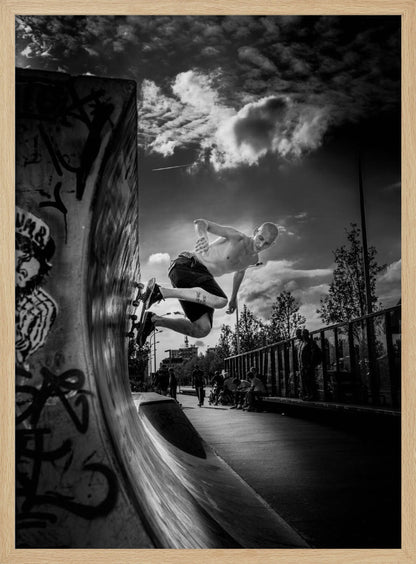 A dramatic black and white, low-angle action shot of a shirtless skateboarder suspended in mid-air at the peak of a graffiti-covered skate ramp, against a backdrop of a cloudy sky. Decor