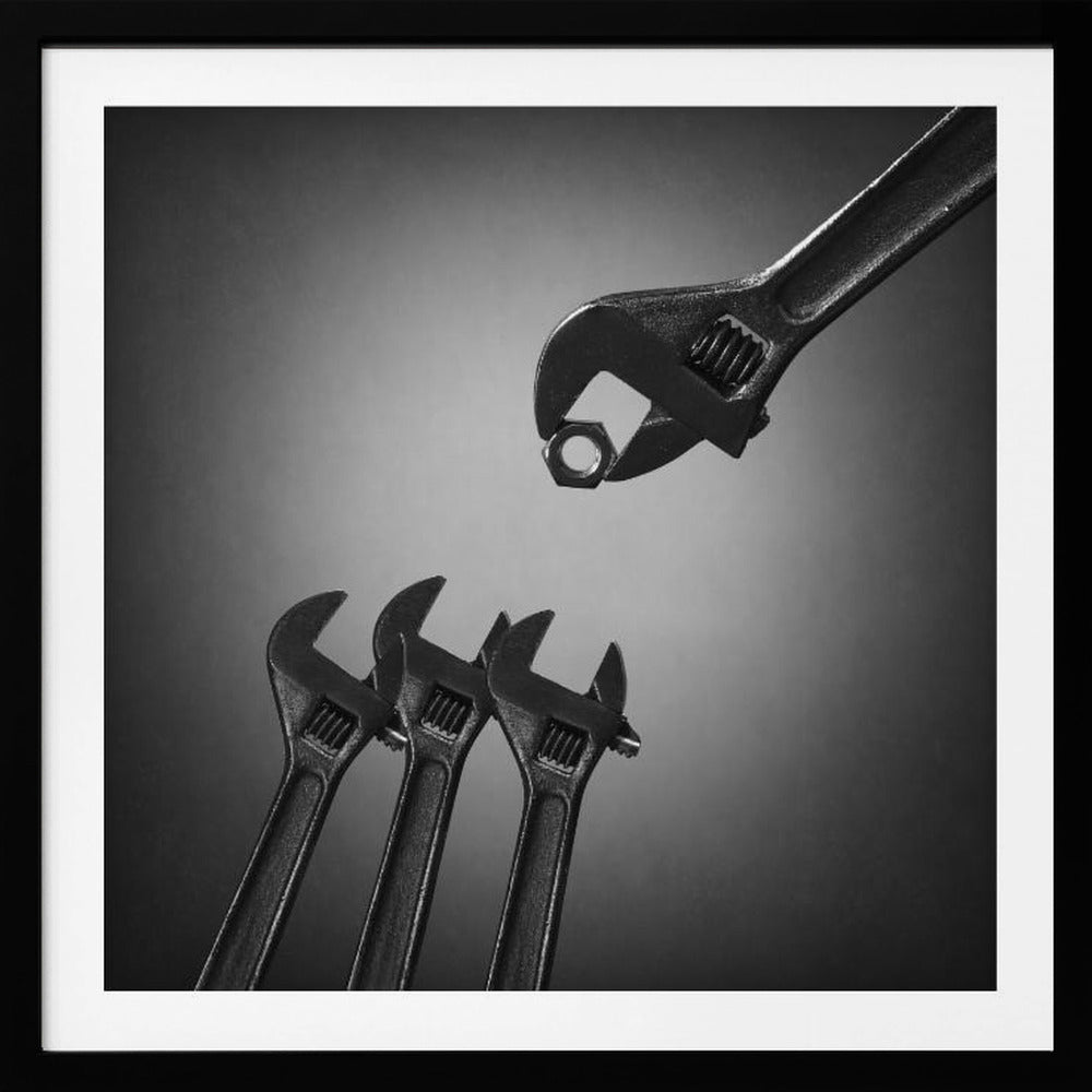 A conceptual black and white photograph in a light wood frame, depicting a large adjustable wrench holding a small nut above three smaller wrenches, which are angled upwards as if they were baby birds waiting to be fed. Print