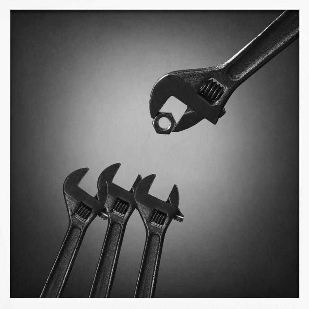 A conceptual black and white photograph in a light wood frame, depicting a large adjustable wrench holding a small nut above three smaller wrenches, which are angled upwards as if they were baby birds waiting to be fed. Print