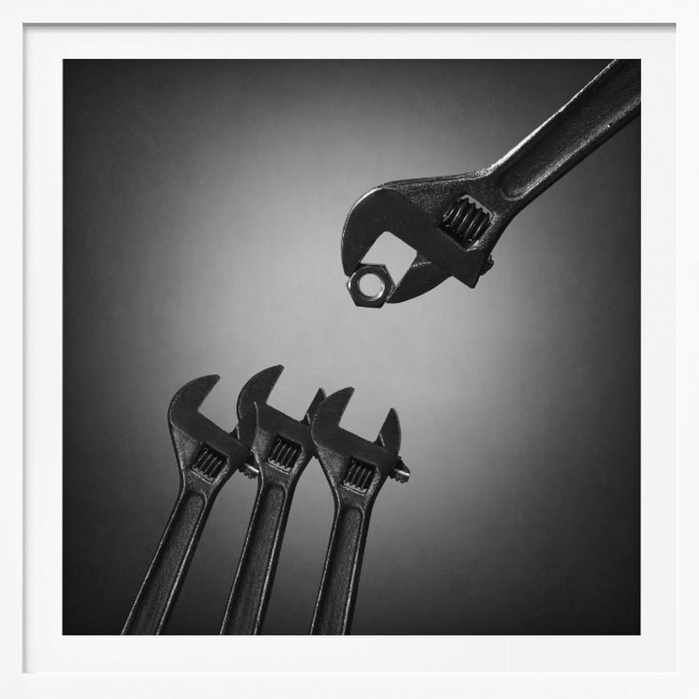 A conceptual black and white photograph in a light wood frame, depicting a large adjustable wrench holding a small nut above three smaller wrenches, which are angled upwards as if they were baby birds waiting to be fed. Print