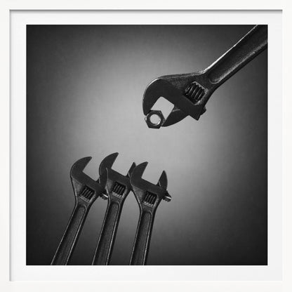 A conceptual black and white photograph in a light wood frame, depicting a large adjustable wrench holding a small nut above three smaller wrenches, which are angled upwards as if they were baby birds waiting to be fed. Print