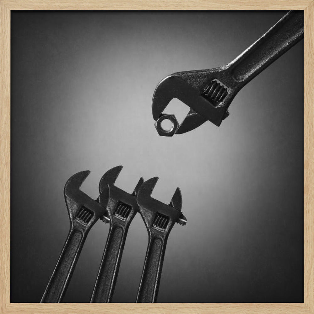 A conceptual black and white photograph in a light wood frame, depicting a large adjustable wrench holding a small nut above three smaller wrenches, which are angled upwards as if they were baby birds waiting to be fed. Print