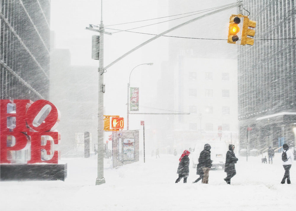 A photograph of a busy city street during a heavy snowstorm. In the foreground on the left, a large, bright red sculpture spells out the word 'HOPE', providing a stark contrast to the muted, white, and gray tones of the snowy scene. Pedestrians in winter coats are crossing the snow-covered street, and tall buildings are partially obscured by the falling snow in the background. Wall Art