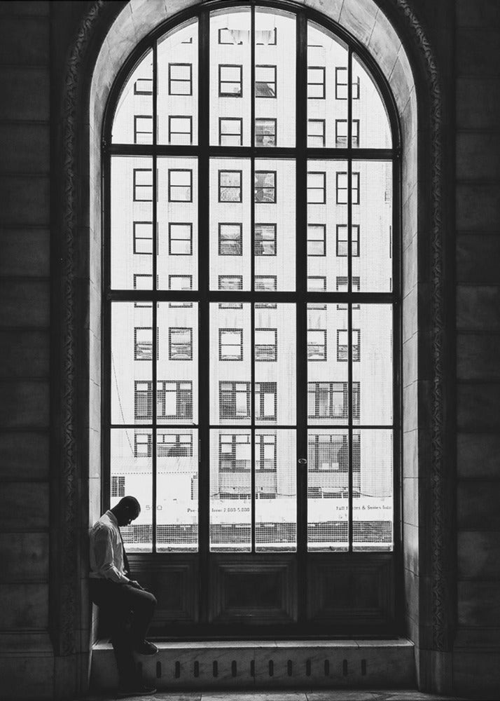 A black and white photograph of a solitary man sitting with his head bowed on the ledge of a massive arched window. The window's dark mullions create a grid over the bright view of another building outside, while the man remains in the shadows, creating a pensive and lonely mood. Poster
