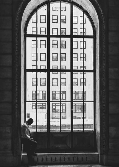 A black and white photograph of a solitary man sitting with his head bowed on the ledge of a massive arched window. The window's dark mullions create a grid over the bright view of another building outside, while the man remains in the shadows, creating a pensive and lonely mood. Poster