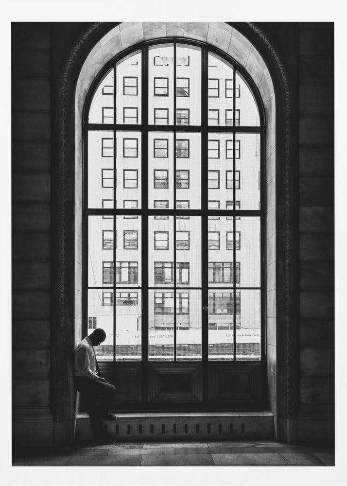 A black and white photograph of a solitary man sitting with his head bowed on the ledge of a massive arched window. The window's dark mullions create a grid over the bright view of another building outside, while the man remains in the shadows, creating a pensive and lonely mood. Poster