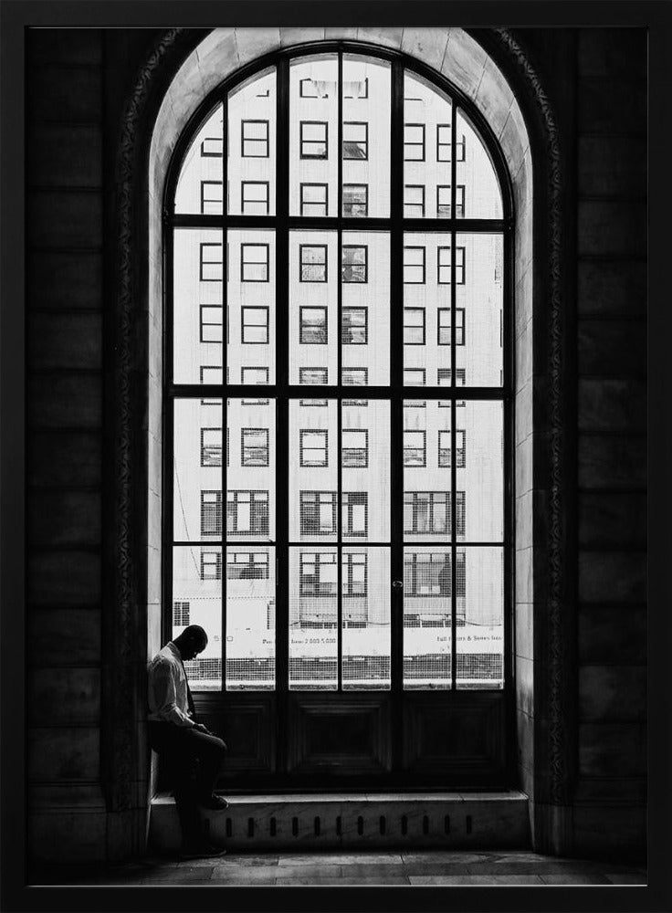 A black and white photograph of a solitary man sitting with his head bowed on the ledge of a massive arched window. The window's dark mullions create a grid over the bright view of another building outside, while the man remains in the shadows, creating a pensive and lonely mood. Poster
