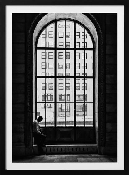 A black and white photograph of a solitary man sitting with his head bowed on the ledge of a massive arched window. The window's dark mullions create a grid over the bright view of another building outside, while the man remains in the shadows, creating a pensive and lonely mood. Poster