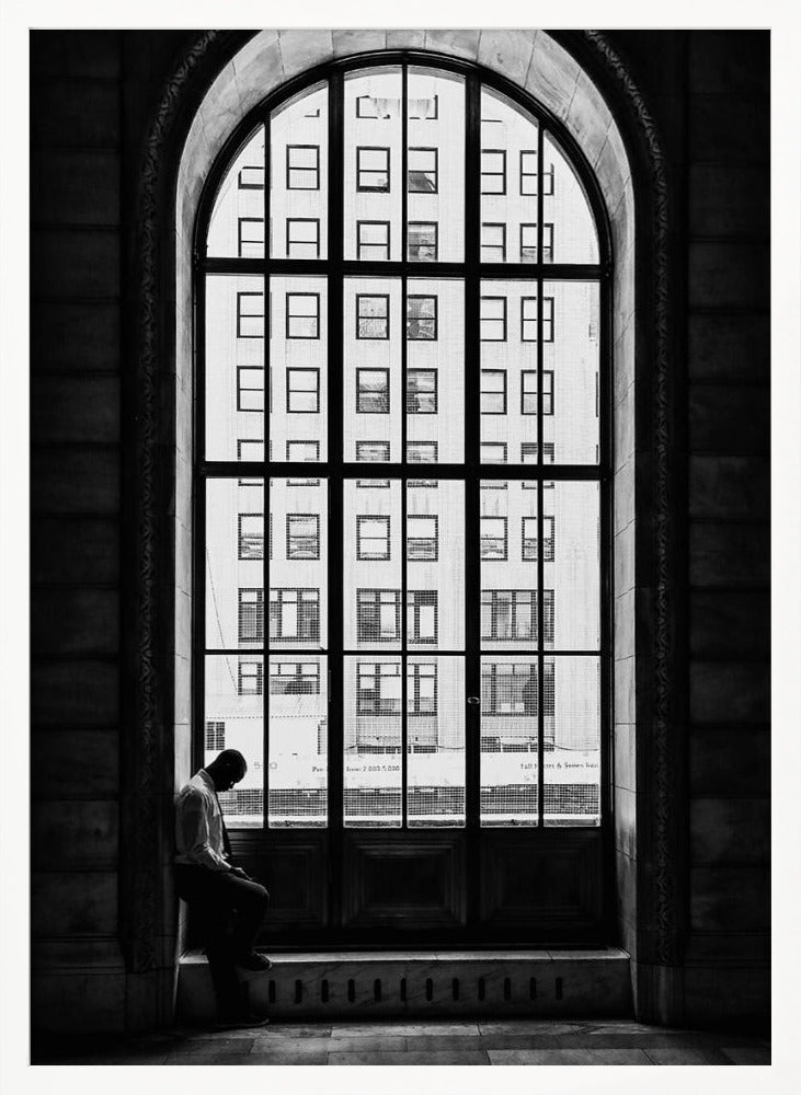 A black and white photograph of a solitary man sitting with his head bowed on the ledge of a massive arched window. The window's dark mullions create a grid over the bright view of another building outside, while the man remains in the shadows, creating a pensive and lonely mood. Poster