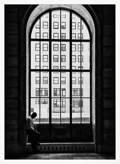 A black and white photograph of a solitary man sitting with his head bowed on the ledge of a massive arched window. The window's dark mullions create a grid over the bright view of another building outside, while the man remains in the shadows, creating a pensive and lonely mood. Poster