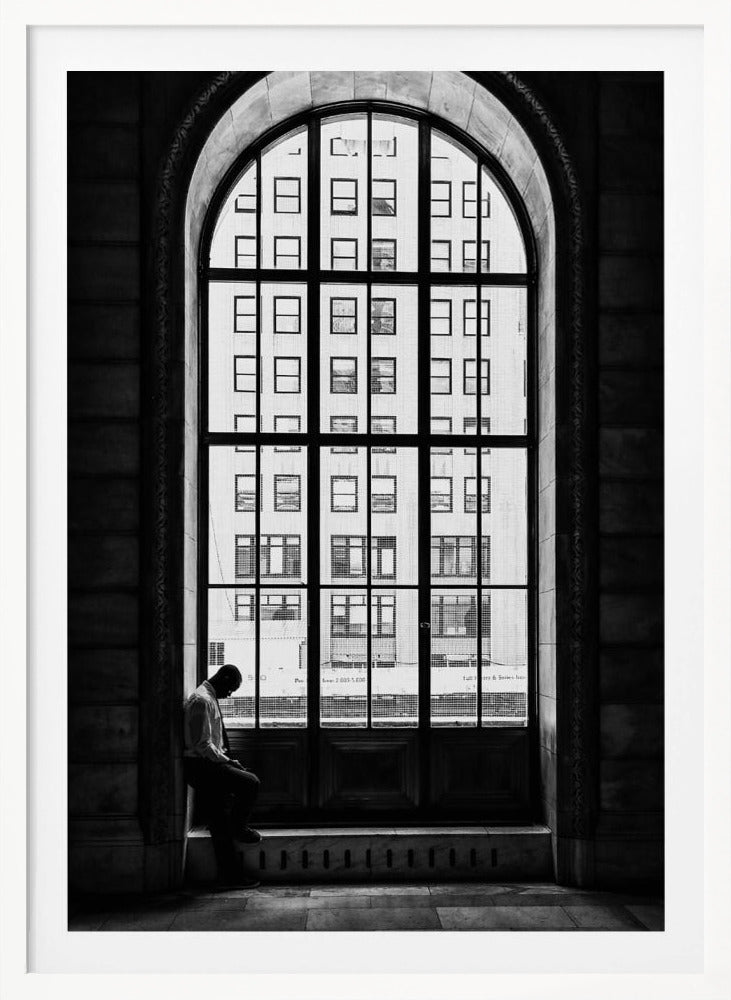 A black and white photograph of a solitary man sitting with his head bowed on the ledge of a massive arched window. The window's dark mullions create a grid over the bright view of another building outside, while the man remains in the shadows, creating a pensive and lonely mood. Poster