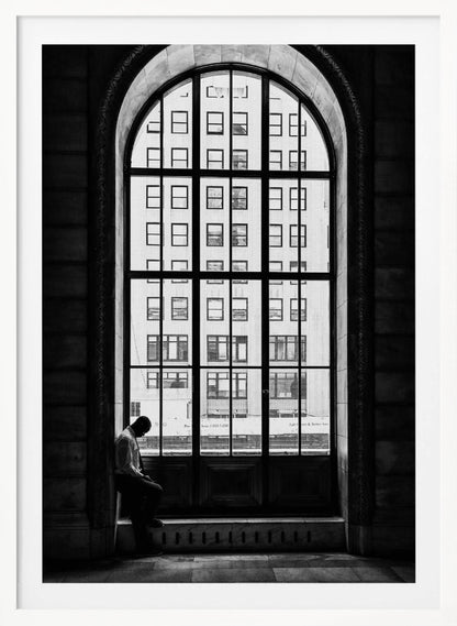 A black and white photograph of a solitary man sitting with his head bowed on the ledge of a massive arched window. The window's dark mullions create a grid over the bright view of another building outside, while the man remains in the shadows, creating a pensive and lonely mood. Poster