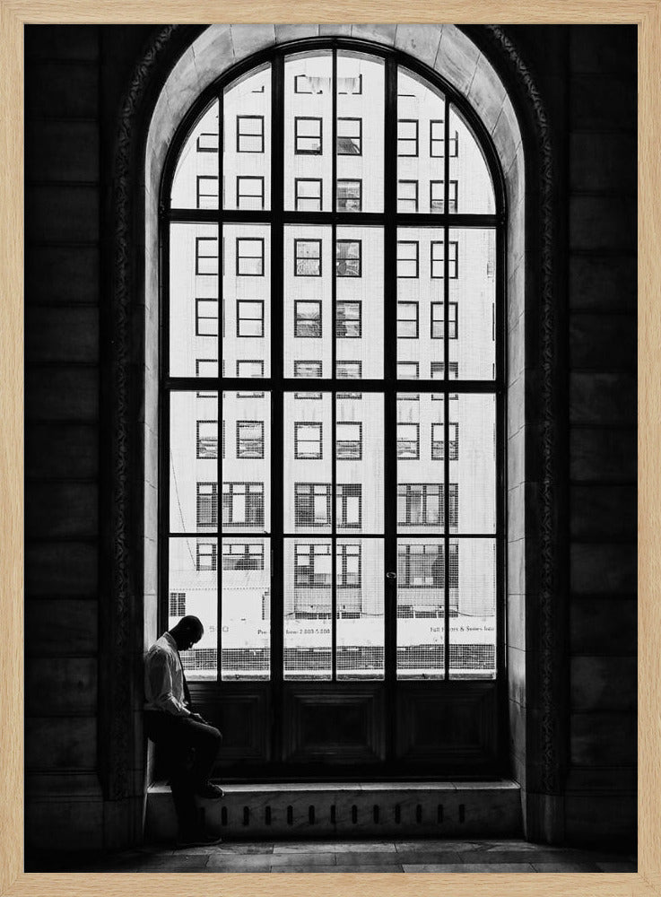 A black and white photograph of a solitary man sitting with his head bowed on the ledge of a massive arched window. The window's dark mullions create a grid over the bright view of another building outside, while the man remains in the shadows, creating a pensive and lonely mood. Poster