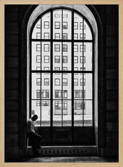 A black and white photograph of a solitary man sitting with his head bowed on the ledge of a massive arched window. The window's dark mullions create a grid over the bright view of another building outside, while the man remains in the shadows, creating a pensive and lonely mood. Poster