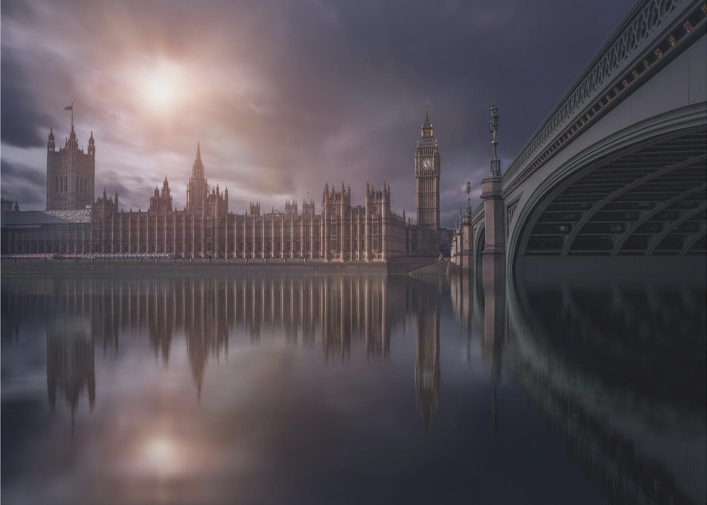 A dramatic, atmospheric photograph of the Houses of Parliament and Big Ben in London, reflected in the still waters of the River Thames. A bright sun breaks through dark, moody clouds, casting a warm light on the scene, with a stone bridge visible to the right. The image is enclosed in a silver frame. Print