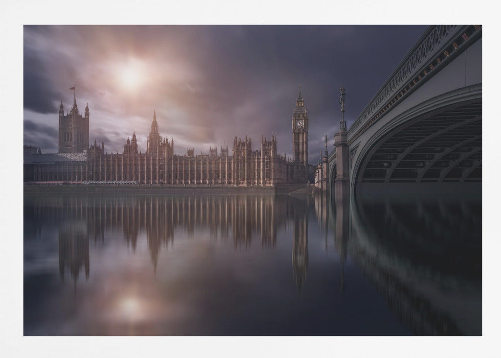 A dramatic, atmospheric photograph of the Houses of Parliament and Big Ben in London, reflected in the still waters of the River Thames. A bright sun breaks through dark, moody clouds, casting a warm light on the scene, with a stone bridge visible to the right. The image is enclosed in a silver frame. Print
