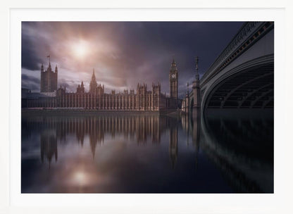A dramatic, atmospheric photograph of the Houses of Parliament and Big Ben in London, reflected in the still waters of the River Thames. A bright sun breaks through dark, moody clouds, casting a warm light on the scene, with a stone bridge visible to the right. The image is enclosed in a silver frame. Print