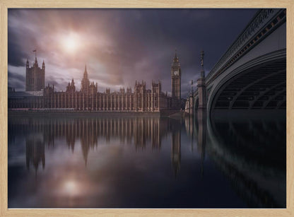 A dramatic, atmospheric photograph of the Houses of Parliament and Big Ben in London, reflected in the still waters of the River Thames. A bright sun breaks through dark, moody clouds, casting a warm light on the scene, with a stone bridge visible to the right. The image is enclosed in a silver frame. Print