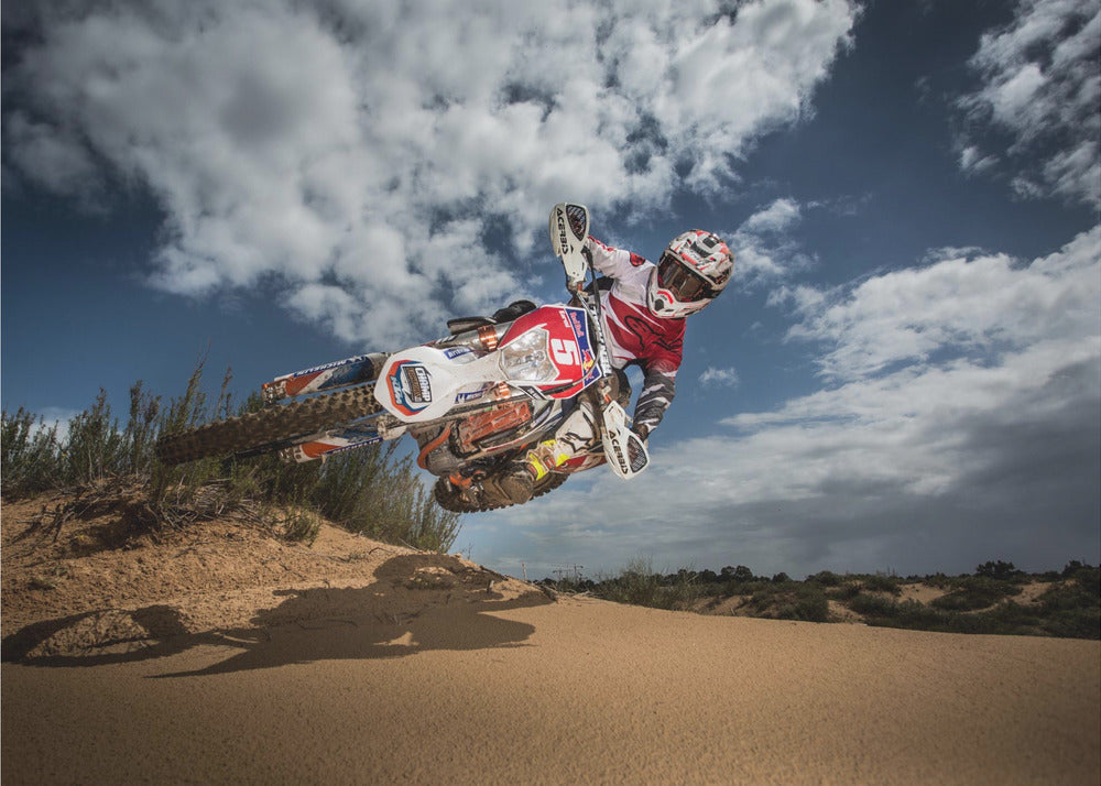 A low-angle action shot of a motocross rider on a dirt bike catching major air over a sand dune against a cloudy blue sky. The image is presented within a silver frame. Decor