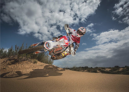 A low-angle action shot of a motocross rider on a dirt bike catching major air over a sand dune against a cloudy blue sky. The image is presented within a silver frame. Decor