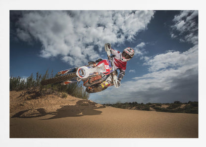 A low-angle action shot of a motocross rider on a dirt bike catching major air over a sand dune against a cloudy blue sky. The image is presented within a silver frame. Decor