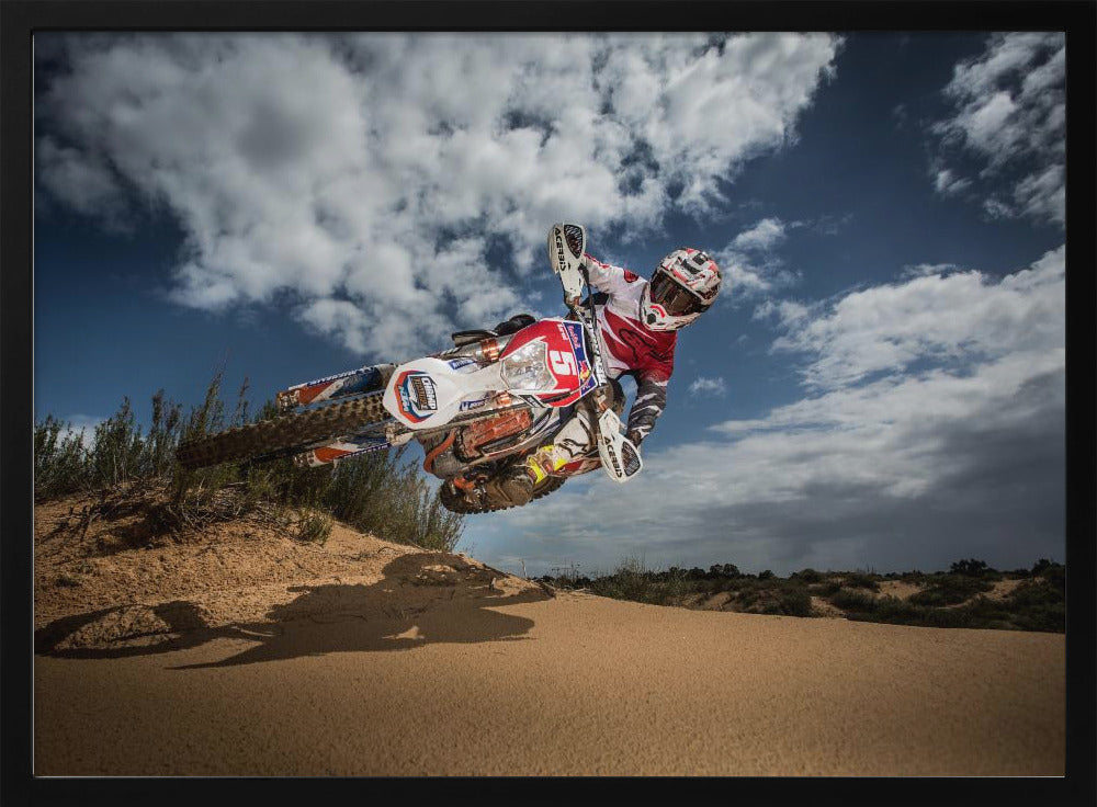 A low-angle action shot of a motocross rider on a dirt bike catching major air over a sand dune against a cloudy blue sky. The image is presented within a silver frame. Decor