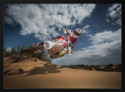A low-angle action shot of a motocross rider on a dirt bike catching major air over a sand dune against a cloudy blue sky. The image is presented within a silver frame. Decor