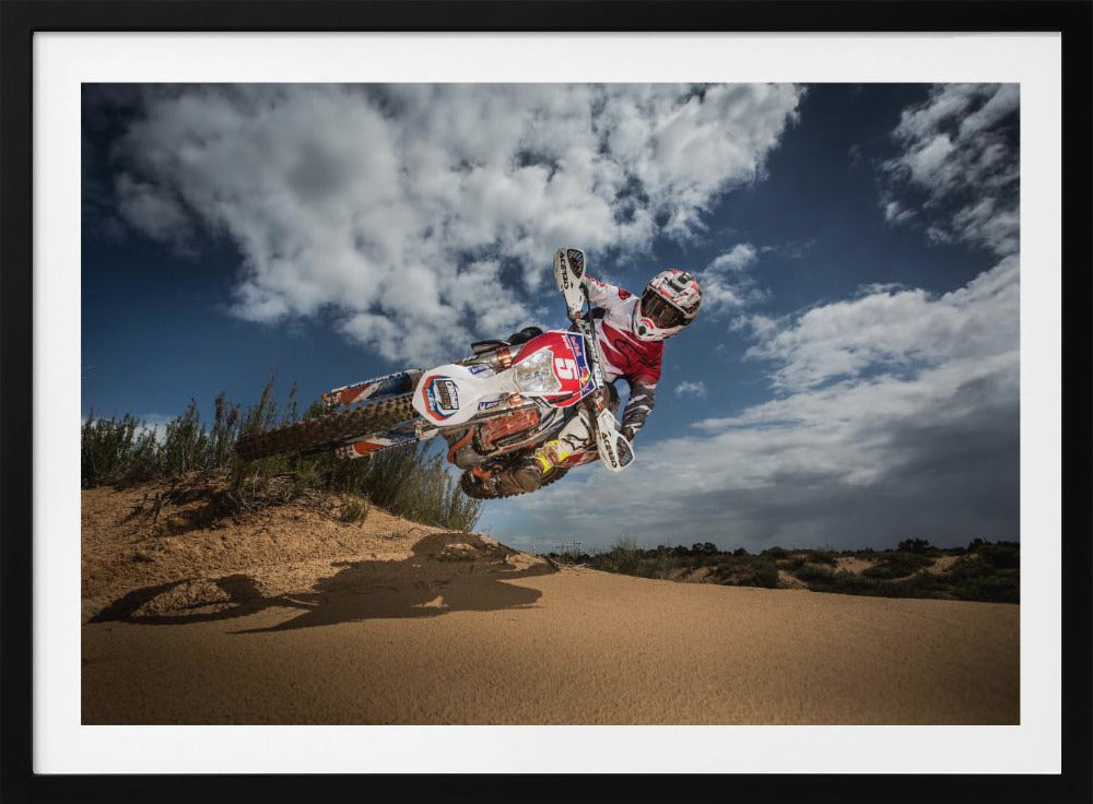A low-angle action shot of a motocross rider on a dirt bike catching major air over a sand dune against a cloudy blue sky. The image is presented within a silver frame. Decor