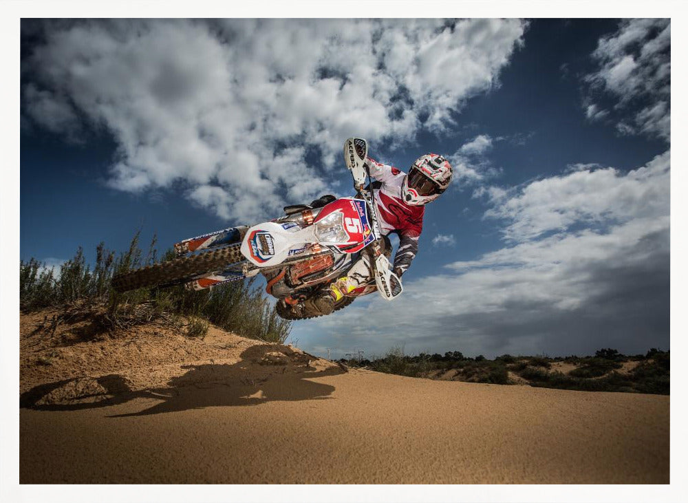 A low-angle action shot of a motocross rider on a dirt bike catching major air over a sand dune against a cloudy blue sky. The image is presented within a silver frame. Decor