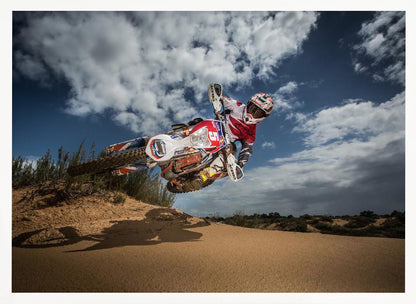 A low-angle action shot of a motocross rider on a dirt bike catching major air over a sand dune against a cloudy blue sky. The image is presented within a silver frame. Decor