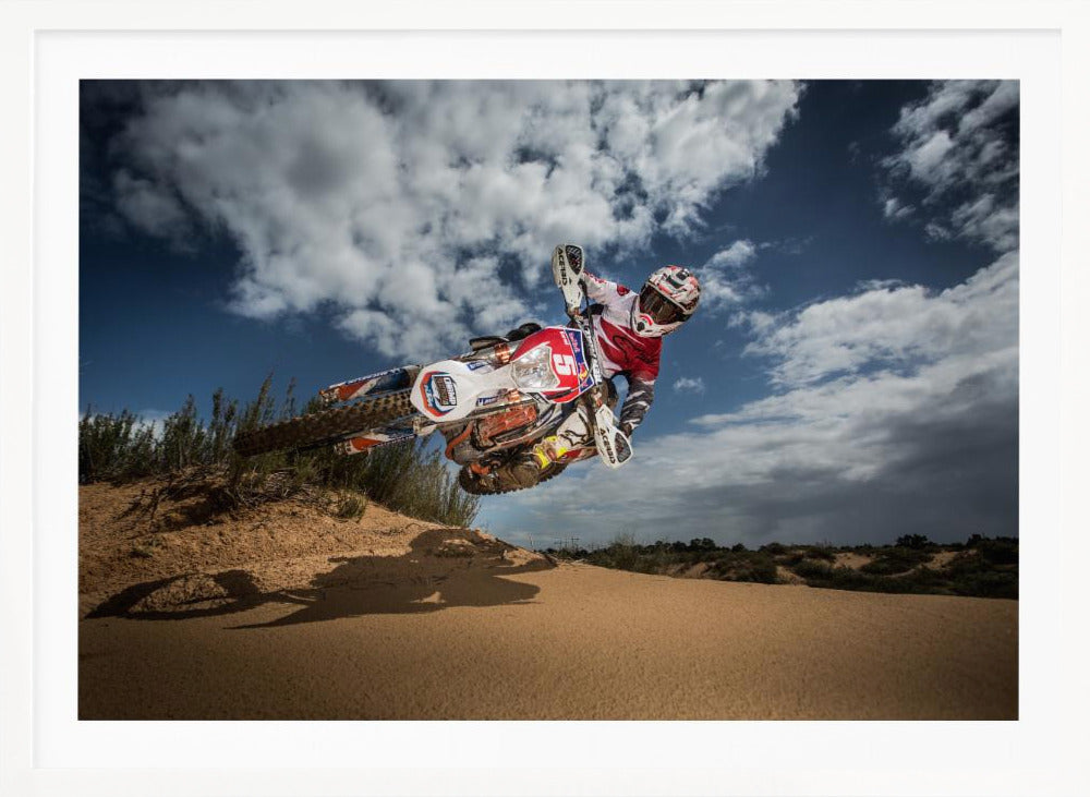 A low-angle action shot of a motocross rider on a dirt bike catching major air over a sand dune against a cloudy blue sky. The image is presented within a silver frame. Decor