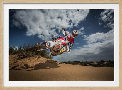 A low-angle action shot of a motocross rider on a dirt bike catching major air over a sand dune against a cloudy blue sky. The image is presented within a silver frame. Decor