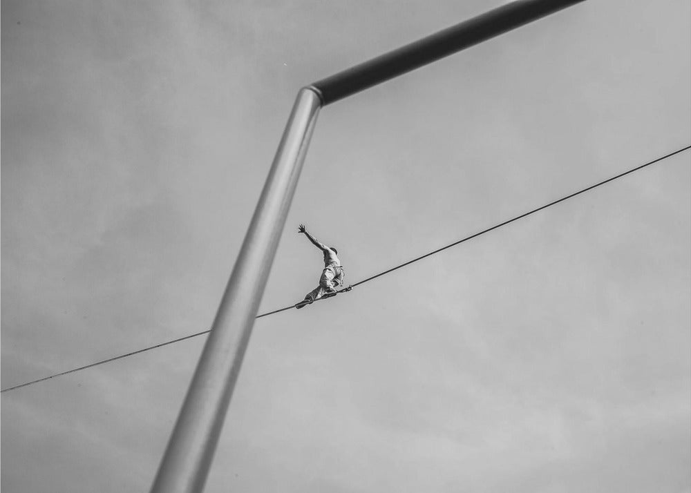 A black and white, low-angle photograph of a person balancing while sitting on a tightrope high in the sky. A large metal pole is in the foreground, creating a strong diagonal line. The image is framed in silver. Decor