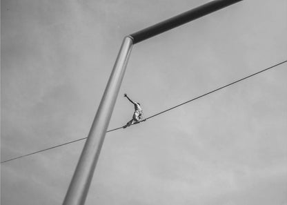 A black and white, low-angle photograph of a person balancing while sitting on a tightrope high in the sky. A large metal pole is in the foreground, creating a strong diagonal line. The image is framed in silver. Decor