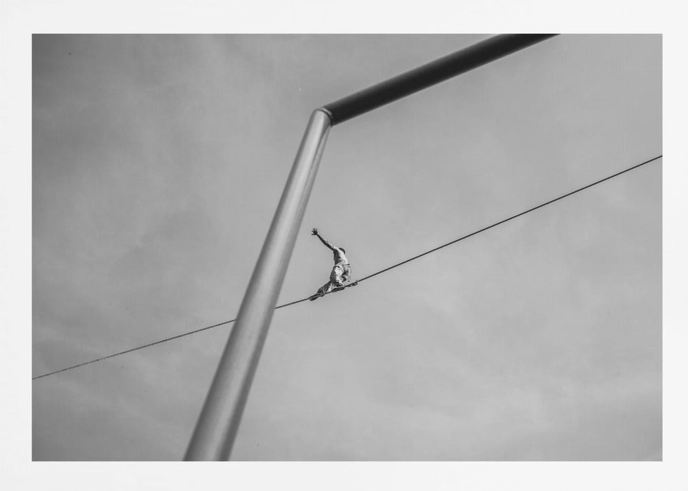 A black and white, low-angle photograph of a person balancing while sitting on a tightrope high in the sky. A large metal pole is in the foreground, creating a strong diagonal line. The image is framed in silver. Decor
