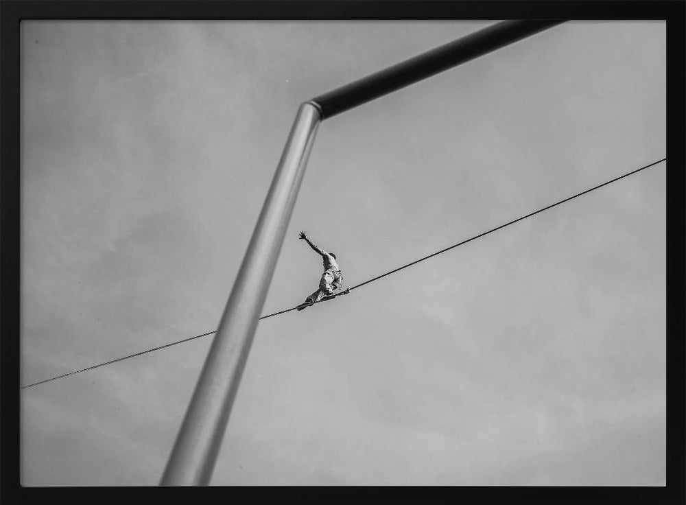 A black and white, low-angle photograph of a person balancing while sitting on a tightrope high in the sky. A large metal pole is in the foreground, creating a strong diagonal line. The image is framed in silver. Decor