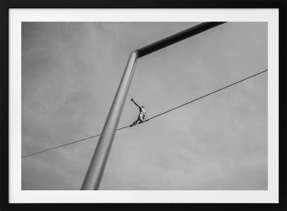 A black and white, low-angle photograph of a person balancing while sitting on a tightrope high in the sky. A large metal pole is in the foreground, creating a strong diagonal line. The image is framed in silver. Decor