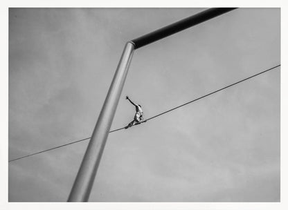 A black and white, low-angle photograph of a person balancing while sitting on a tightrope high in the sky. A large metal pole is in the foreground, creating a strong diagonal line. The image is framed in silver. Decor