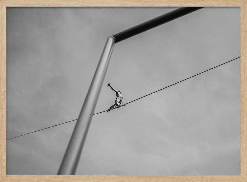 A black and white, low-angle photograph of a person balancing while sitting on a tightrope high in the sky. A large metal pole is in the foreground, creating a strong diagonal line. The image is framed in silver. Decor