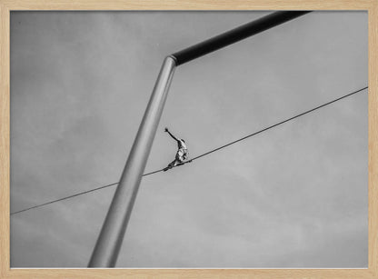 A black and white, low-angle photograph of a person balancing while sitting on a tightrope high in the sky. A large metal pole is in the foreground, creating a strong diagonal line. The image is framed in silver. Decor